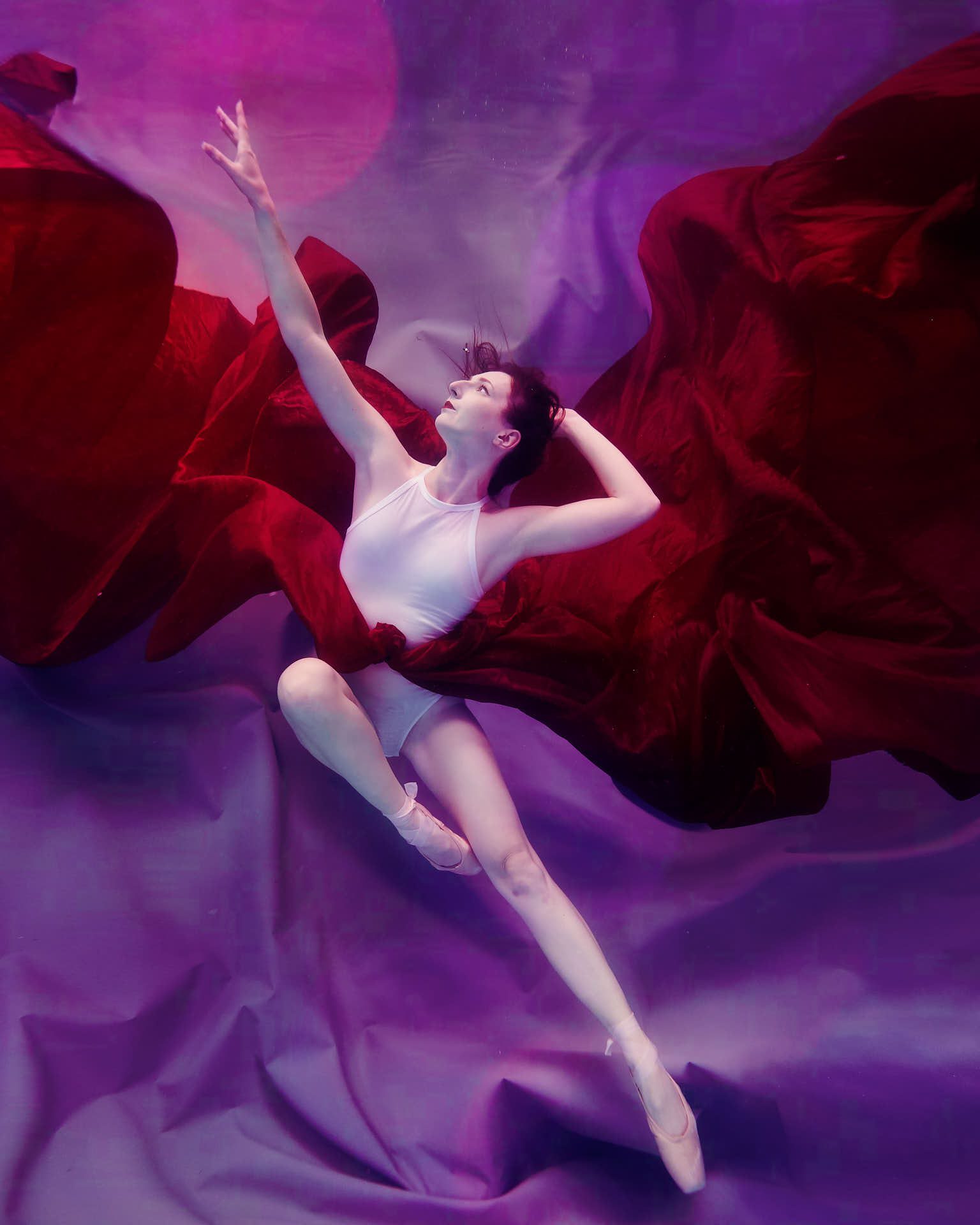 Underwater ballet dancer wearing a white bodysuit against a purple backdrop, with red fabric flowing